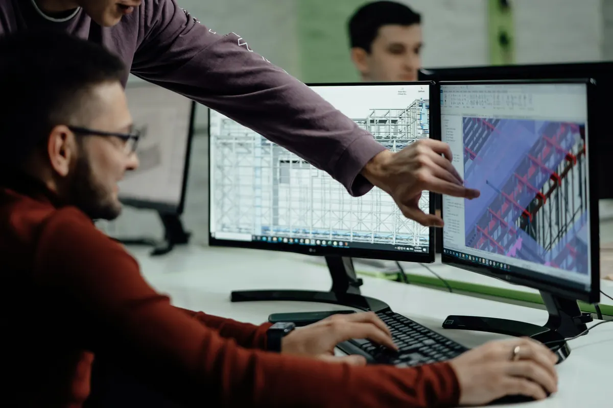 Duas pessoas diante de monitores com o modelo tridimensional estrutural de um edifício aberto em software de projeto, uma delas apontando para a tela
