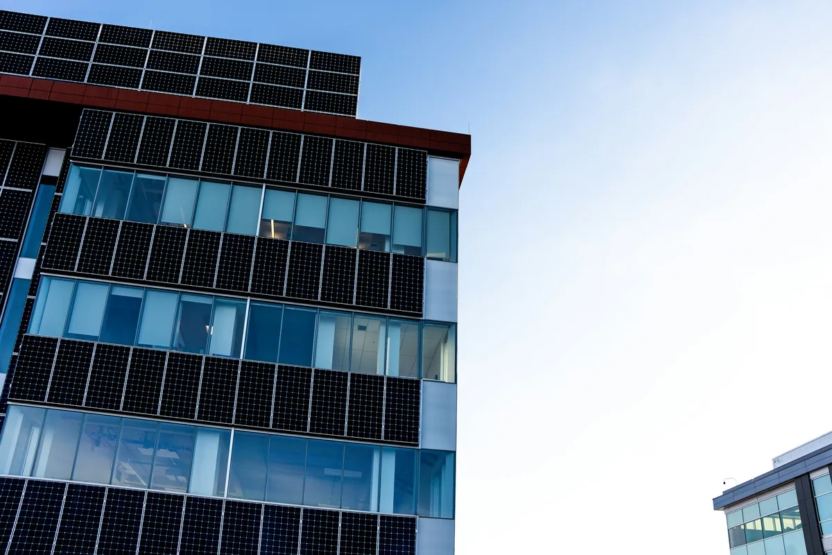 Fachada de edifício moderno revestida por painéis fotovoltaicos alternados com faixas de vidro, vista de baixo sob céu azul